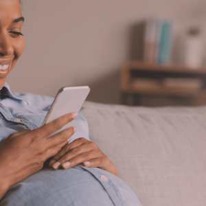 Pregnant woman sitting on a couch, looking at her smarphone and smiling.